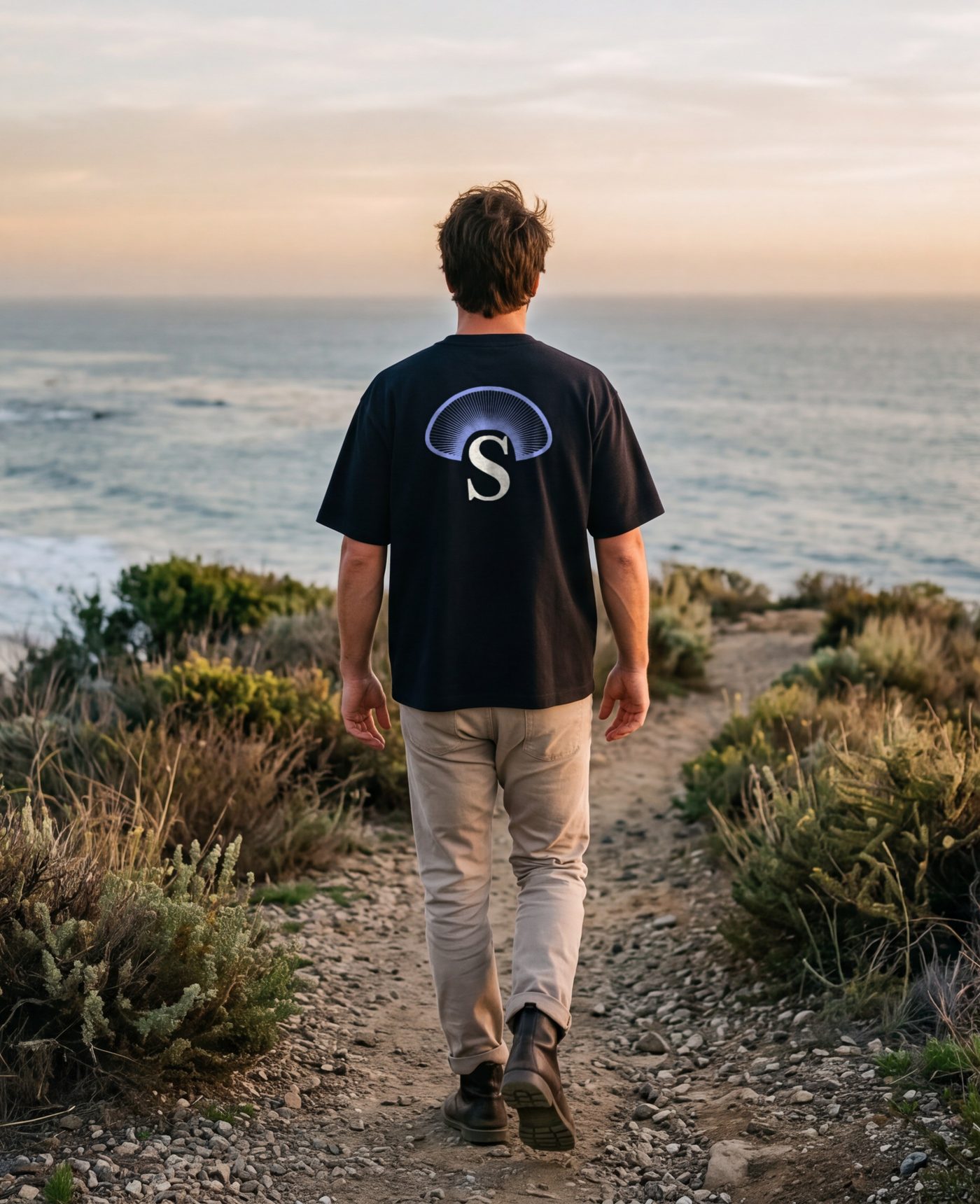 A man walking the coast path above the beach in Malibu, the Cap of Gills across the back of his near-black tee.